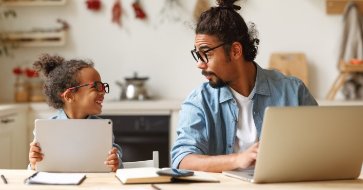 Father using laptop while sitting next to child using a tablet.